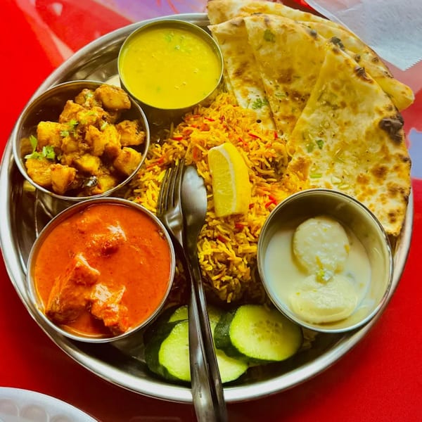 A Bombay Kitchen thali with biryani, naan, curries, and dessert on a red tablecloth.